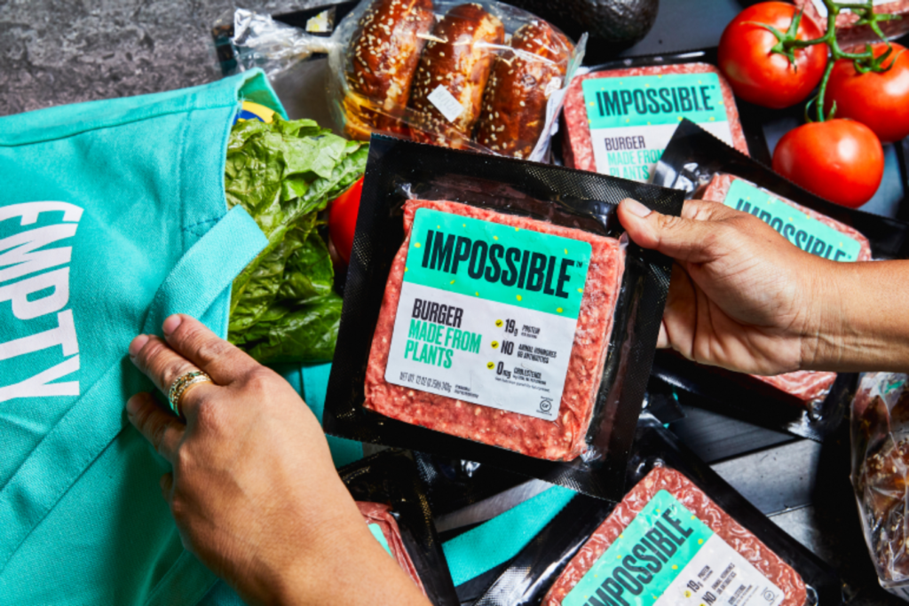 Zwei Hände packen eine Fleischpackung mit der Aufschrift "Impossible" in eine Tüte. Darin sind bereits andere Lebensmittel.
